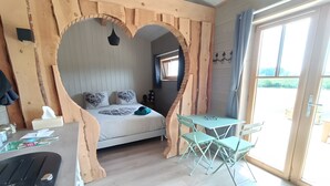 A room with a heart-shaped arch, a bed with patterned pillows, a small table with chairs, and a view through a glass door. - Cabin “Robin Hood’s Cabin” with Private Terrace and Nordic Bath (Foulcrey)