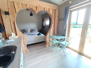 Une piĂšce dotĂ©e dâune arche en forme de cĆur, dâun lit agrĂ©mentĂ© dâoreillers Ă motifs, dâune petite table avec des chaises et dâune vue sur lâextĂ©rieur, visible Ă travers une porte vitrĂ©e.