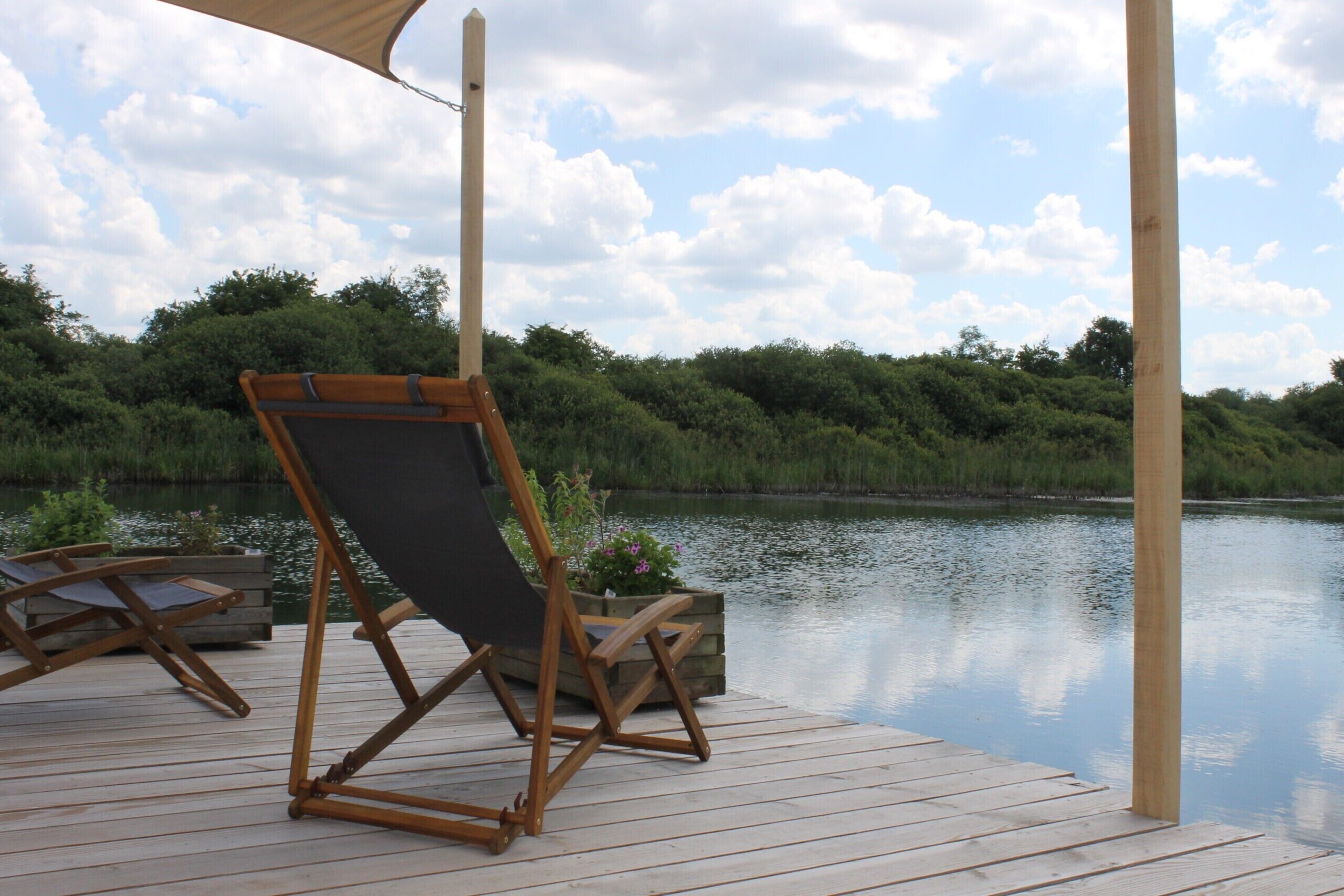 Une terrasse en bois avec un chevalet en toile et une chaise, donnant sur un lac paisible, avec une végétation luxuriante en arrière-plan.