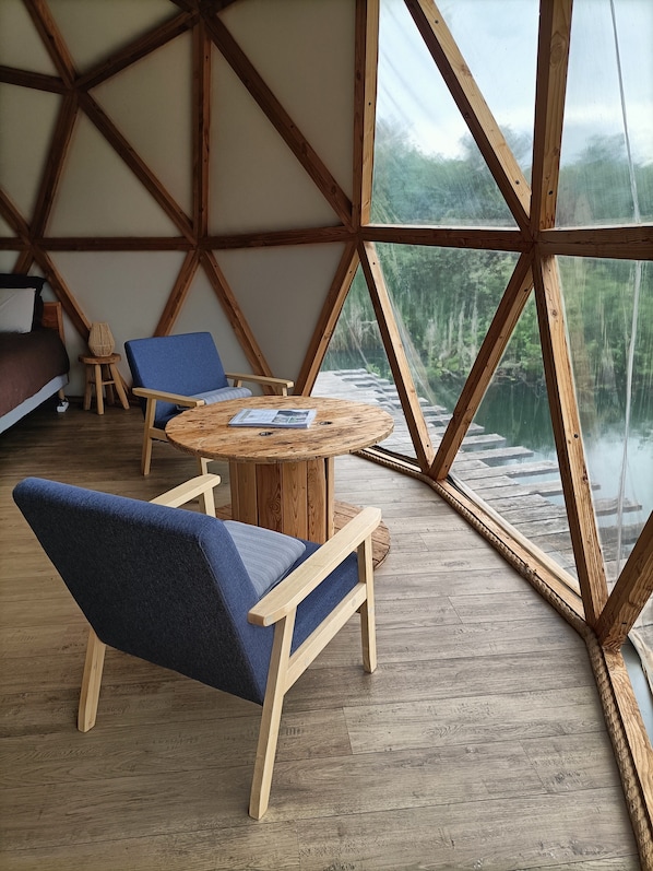 A room with a wooden ceiling structure, a round wooden table, two blue chairs, and a view of a forest through large windows.