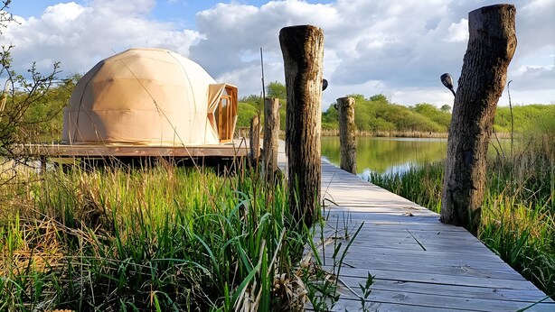 Une structure en forme de dôme, dotée d’une porte en bois, d’un quai en bois et bordée par une étendue d’eau.