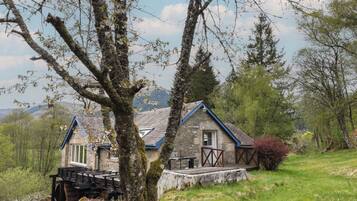 A stone house with a wooden deck, surrounded by trees and a grassy hill.