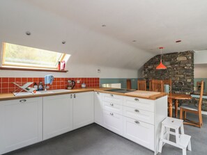 A kitchen with white cabinets, a wooden countertop, a stone wall, and a dining area with wooden chairs and a table. - Culearn Mill (Lochearnhead)