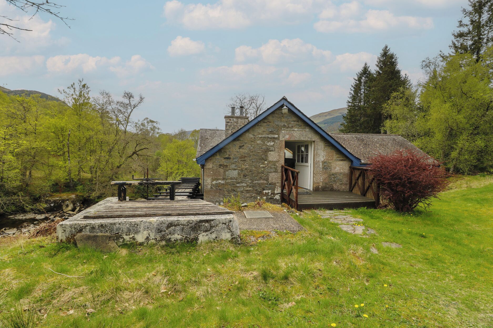 A stone cottage with a wooden deck, a bench, and a small table.