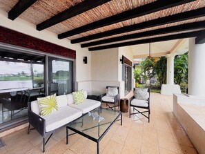 Une terrasse couverte avec une table basse en verre, des canapés blancs ornés de coussins à motifs et des fauteuils en osier.