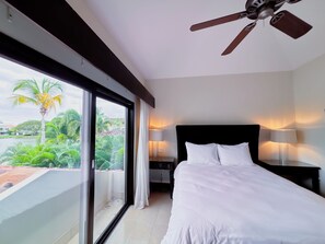 Une chambre d’hôtel avec un lit, deux lampes de chevet, un ventilateur de plafond et une grande fenêtre donnant sur un espace verdoyant et un immeuble.