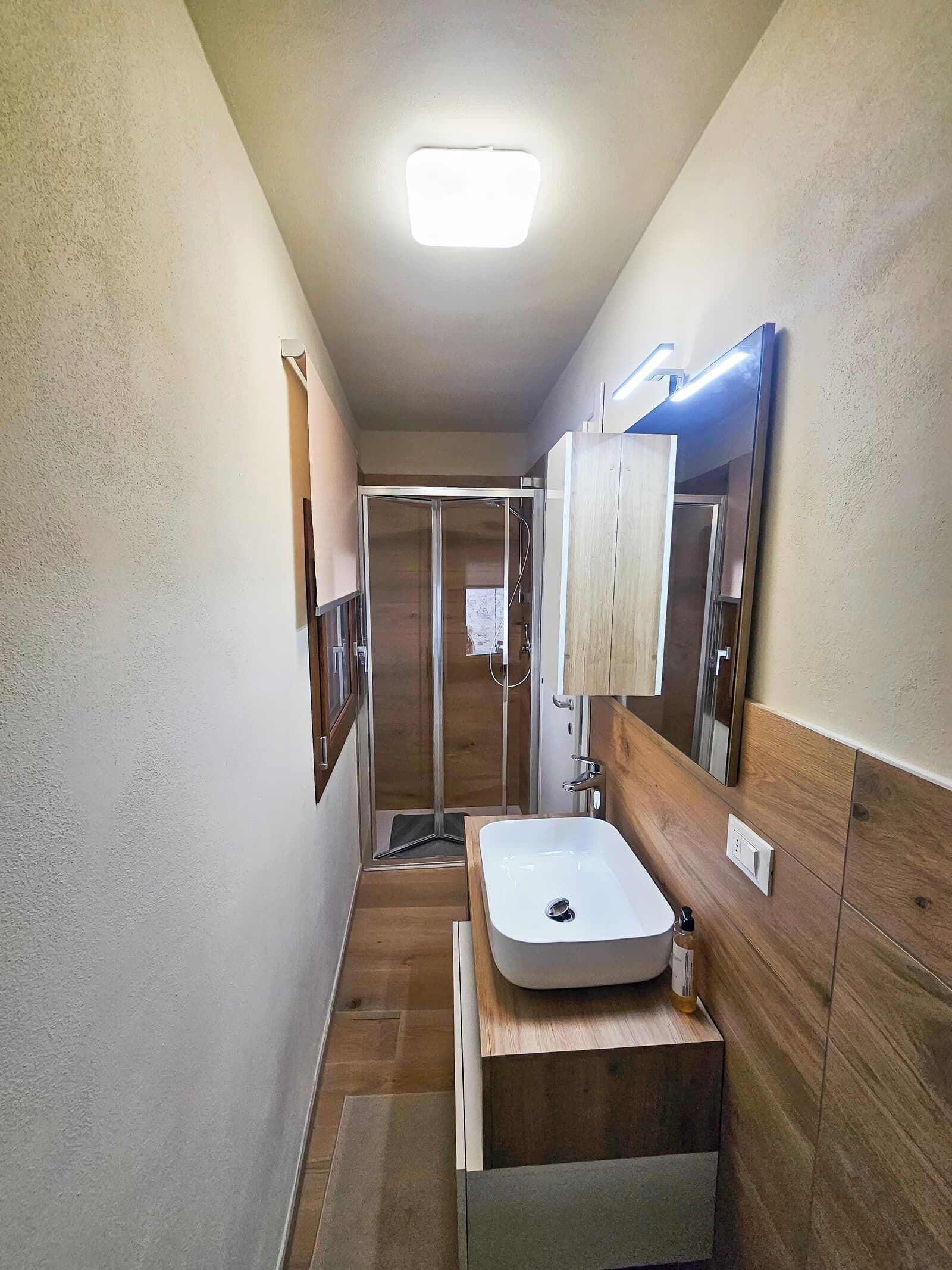Een smalle badkamer met een witte wastafel, houten opbergmeubel en een douchecabine met glazen wanden.
