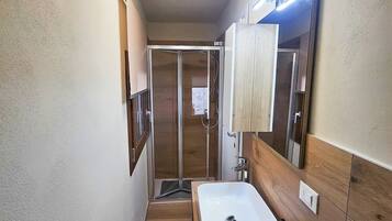 Een smalle badkamer met een witte wastafel, houten opbergmeubel en een douchecabine met glazen wanden.