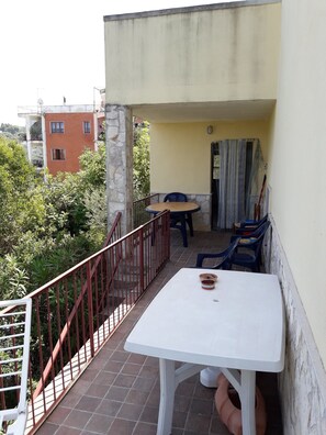 Ein Balkon mit einem weißen Tisch und blauen Stühlen, einem roten Geländer sowie Blick auf Grünflächen und Gebäude.