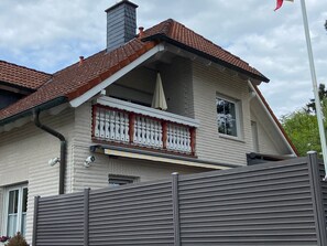 Una casa de dos pisos con techo de tejas rojas, un balcón con barandillas blancas y una chimenea.
