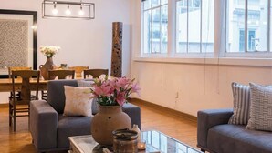 Ein modernes Wohnzimmer mit einem Glastisch, zwei Sofas, einer Essecke mit Holztisch und Stühlen und großen Fenstern mit Blick auf die Stadt.
