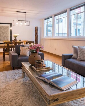 A modern living room with a glass coffee table, two sofas, a dining area with a wooden table and chairs, and large windows offering a view of the city. - Location- Design and Comfort Fully Equipped (Mexico City)