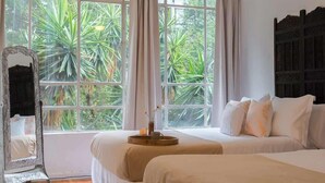 Ein Schlafzimmer mit zwei Betten, einem Deckenventilator, einem großen Fenster mit Vorhängen und Blick ins Grüne.