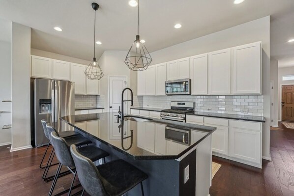 A modern kitchen with white cabinets, a stainless steel refrigerator, a built-in oven, and a large island with bar stools. - Adorable 3-bedroom Basecamp ATX (Buda)