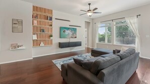 A living room with a sectional sofa, a wooden bookshelf, a mounted TV, and a ceiling fan. - Adorable 3-bedroom Basecamp ATX (Buda)