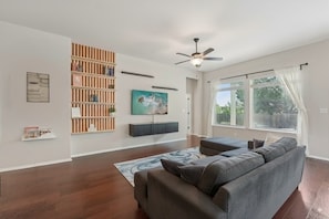 A living room with a sectional sofa, a wooden bookshelf, a mounted TV, and a ceiling fan. - Adorable 3-bedroom Basecamp ATX (Buda)