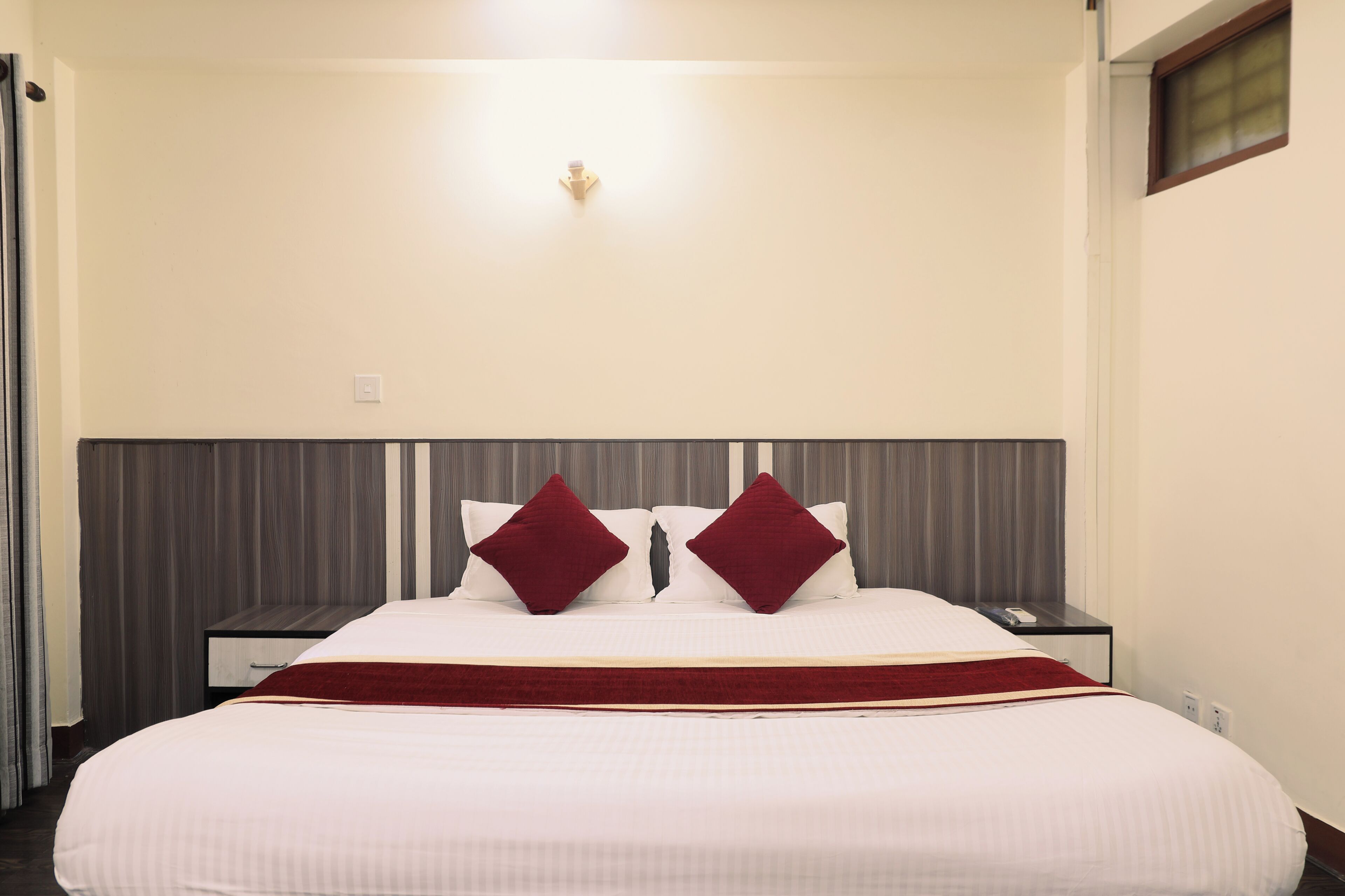 A hotel room with a large bed, two red pillows, a wooden headboard, a bedside table with a phone, and a red throw blanket.