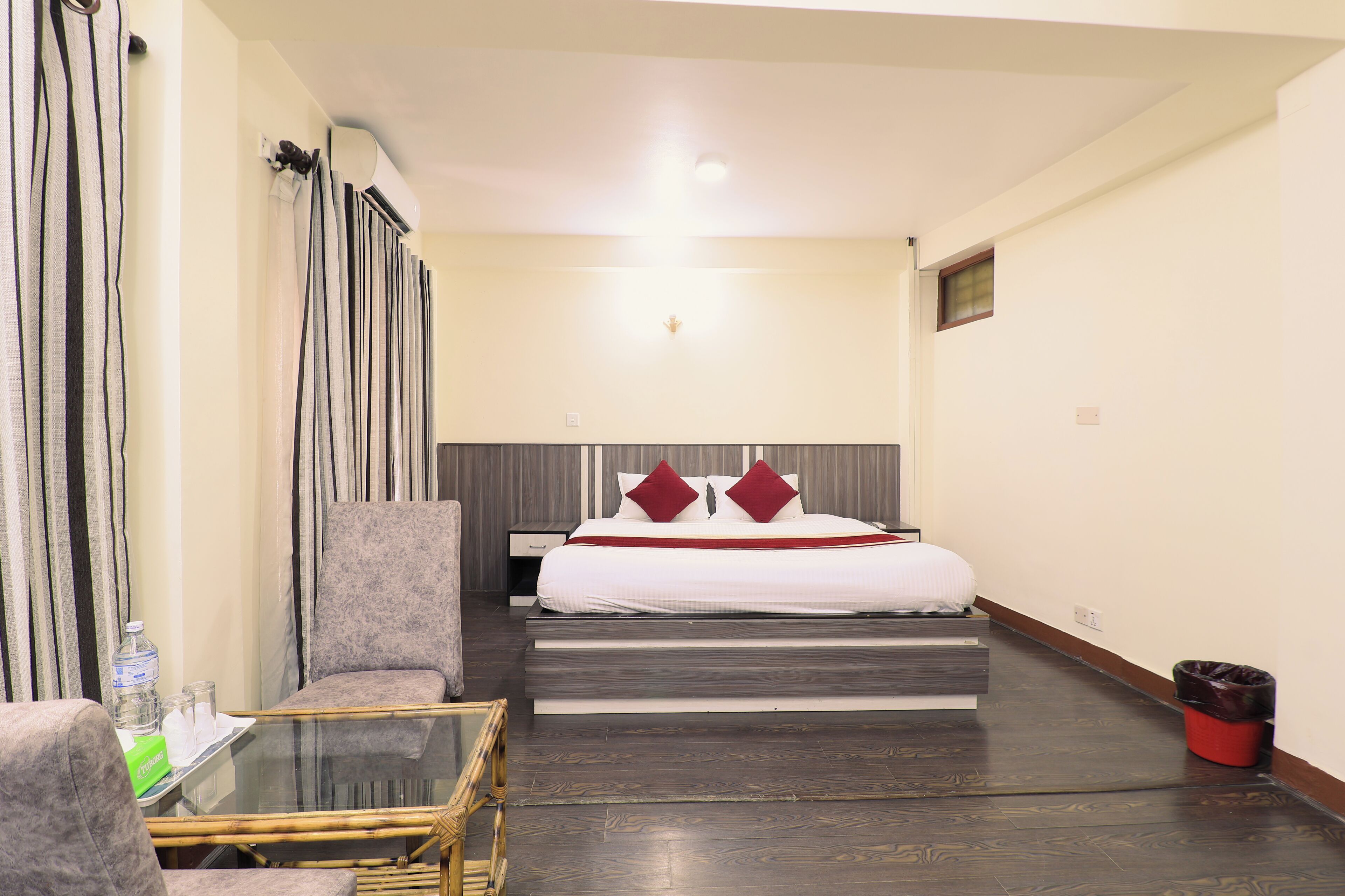 A hotel room with a bed, a chair, a glass table, and a trash bin.