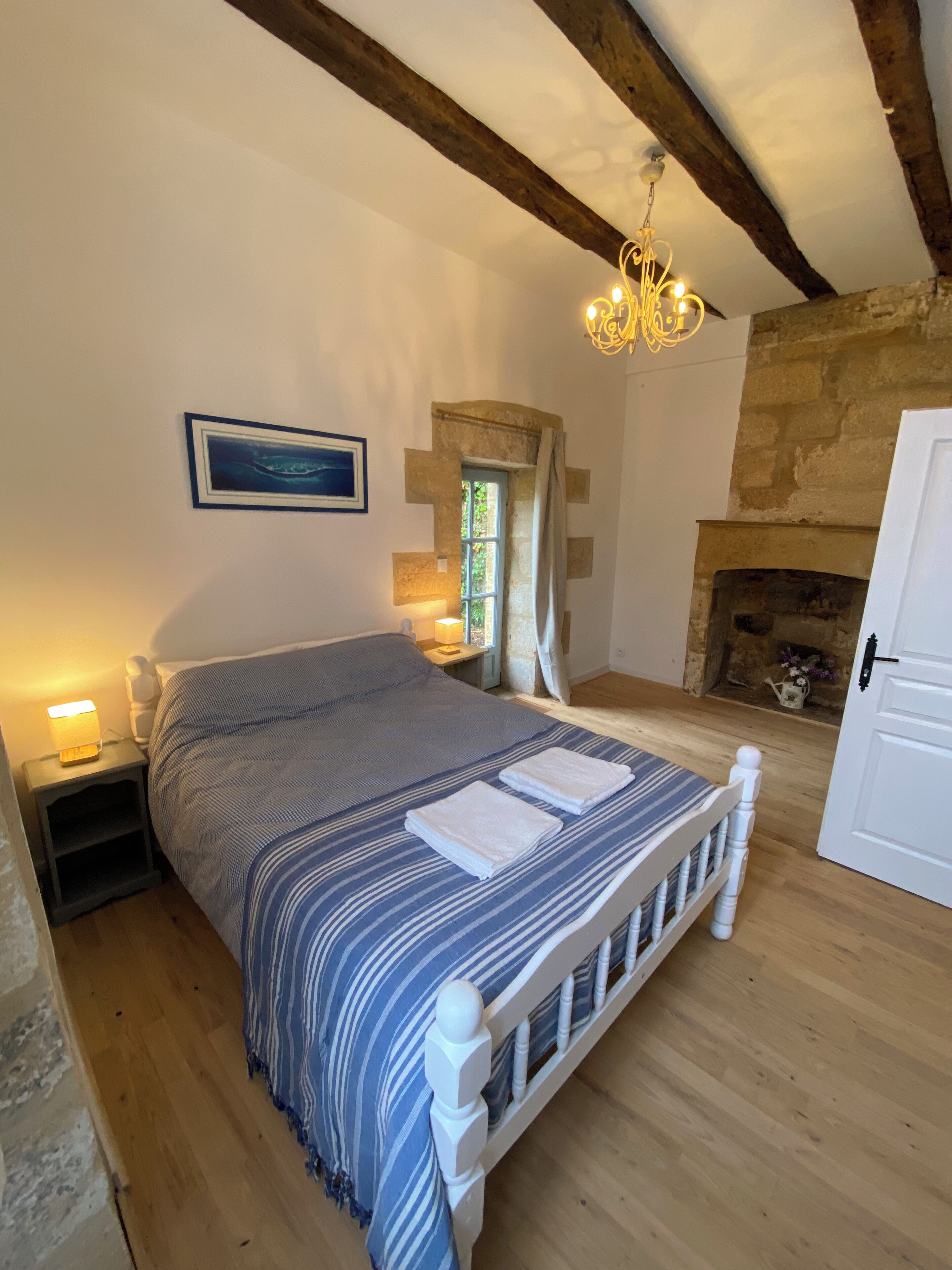 A bedroom with a wooden bed, a nightstand, a wall-mounted picture, a stone fireplace, and a chandelier.