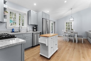 A modern kitchen with grey cabinets, a stainless steel refrigerator, a coffee maker, and a dining area with a table and chairs. - The Retreat at Seal Point-New! (Lamoine)