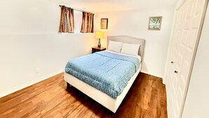 A bedroom with a bed, a nightstand, a lamp, a window with curtains, and a door. - Elegant 1BR Retreat Near Downtown Relax in Comfort (Dieppe)