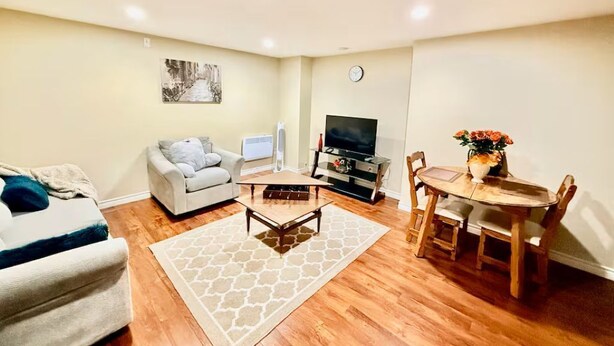 A living room with a sofa, a coffee table, a TV, a dining table with chairs, and a clock on the wall. - Elegant 1BR Retreat Near Downtown Relax in Comfort (Dieppe)
