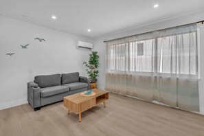 Ein modernes Wohnzimmer mit einem grauen Sofa, einem Holztisch und einem großen Fenster mit leichten Vorhängen.