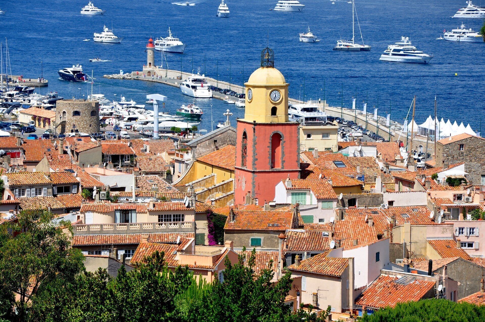 Une ville côtière qui se distingue par une tour jaune et rouge imposante, de nombreux bateaux dans le port et des bâtiments traditionnels aux toits de tuiles rouges.