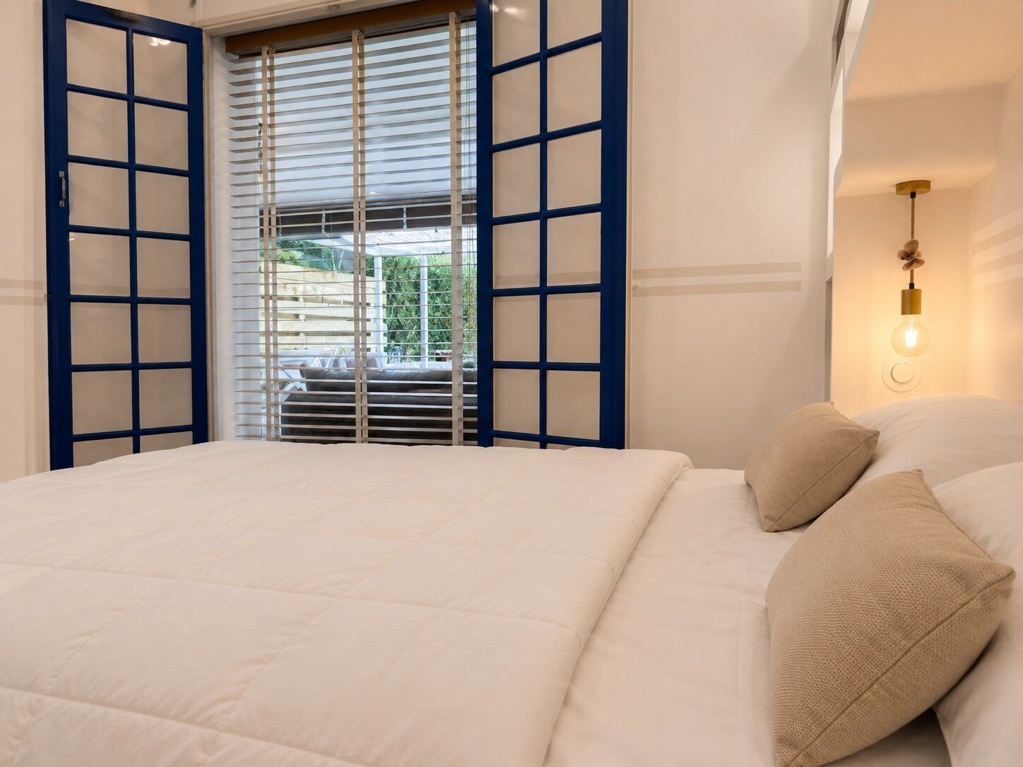 A bedroom with a bed, a window with blinds, a wall-mounted light fixture, and a view of greenery outside.
