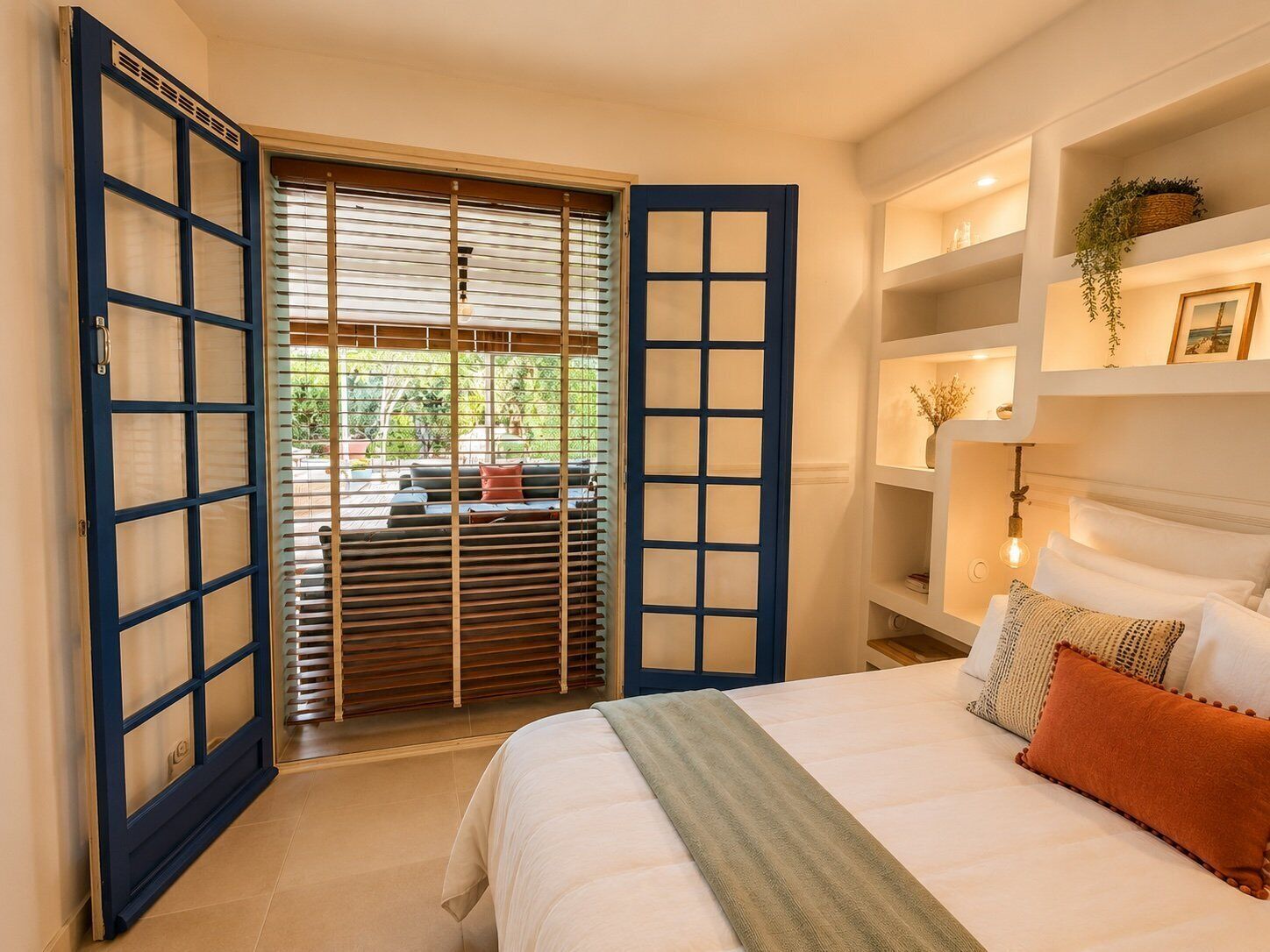 Une chambre à coucher avec un lit, une fenêtre avec des stores, une étagère avec des objets décoratifs et une vue sur l’extérieur.