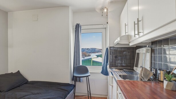 Una habitación pequeña con cama, un taburete, ventana con vistas, kitchenette con lavabo y cocina, e iluminación en el techo.