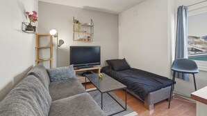Una sala moderna con un sofá, una mesita, un televisor y una cama. Hay estanterías con plantas y una vista de agua a través de una ventana.