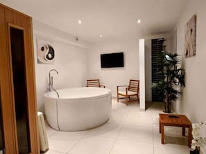 Een moderne badkamer met een vrijstaand ligbad, twee houten stoelen, een klein houten tafeltje en een potplant.