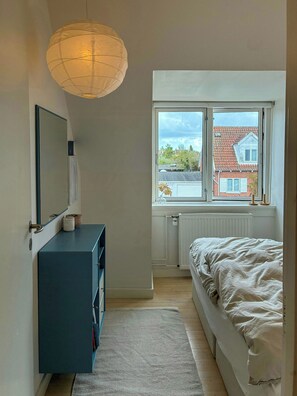 Et soveværelse med en seng, et kommode, et spejl, et vindue med udsigt til bygninger, og en hængende lampe.