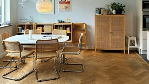A dining area with a round table, wicker chairs, a hanging light fixture, and a cabinet with a plant. - Townhouse with art and terrace (København)