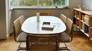 A dining area with a round table, six wicker chairs, a hanging pendant light, a bookshelf, and a window with a view of buildings and greenery. - Townhouse with art and terrace (København)