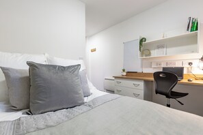 Ein Schlafzimmer mit Bett, Kissen, Schreibtisch mit Computer, Stuhl und Regal mit Büchern und Pflanze.