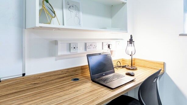 Ein Laptop auf einem Holztisch, zusammen mit einem schwarzen Stuhl, einer Schreibtischlampe und einem Regal mit Pflanze und gerahmten Kunstwerken.