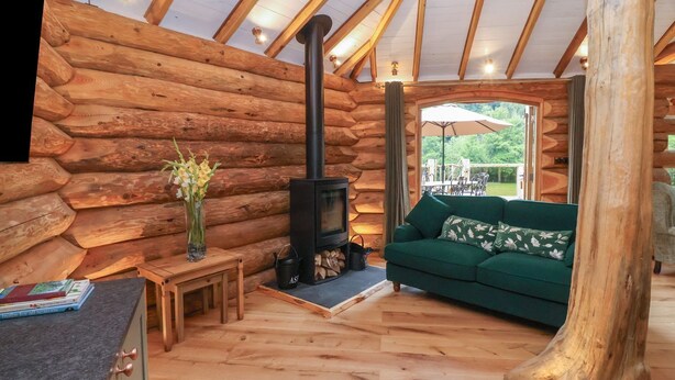 A log cabin interior with a fireplace, a sofa, a dining area, and a view of the outdoors.