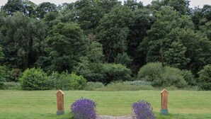 A stone archway with two wooden posts on either side, surrounded by a grassy field and dense trees in the background.