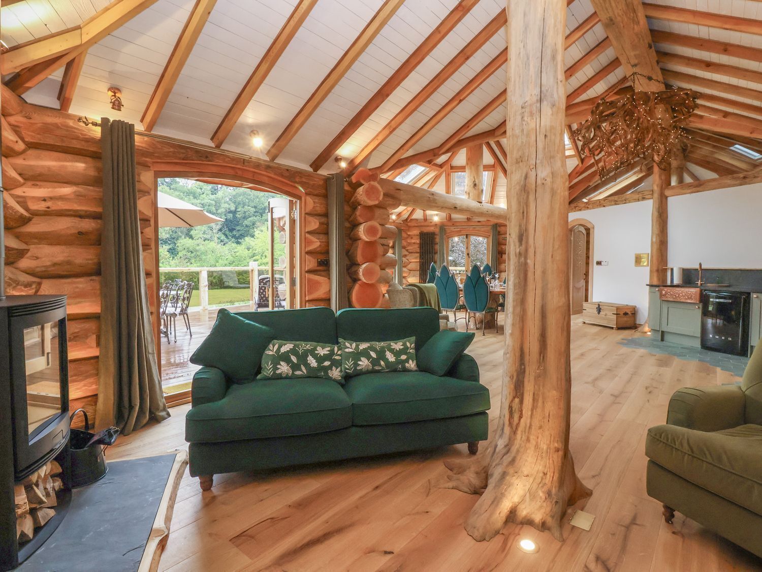 Interior de una cabaña de troncos con chimenea, un sofá verde, piso de madera y un área de comedor visible a través de una puerta abierta.