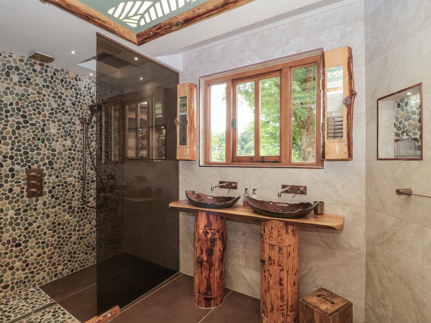 A bathroom with a stone mosaic wall, a wooden vanity with a large window, and a glass shower enclosure.