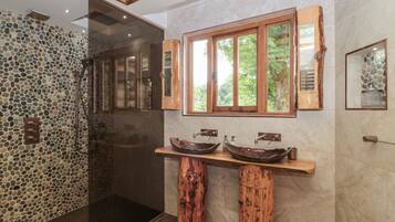 A bathroom with a stone mosaic wall, a wooden vanity with a large window, and a glass shower enclosure.