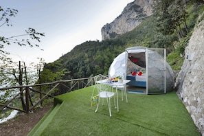 Ein gläsernes, kuppelförmiges Zimmer mit Bett, Tisch und Stuhl, auf künstlichem Gras mit Bergblick.