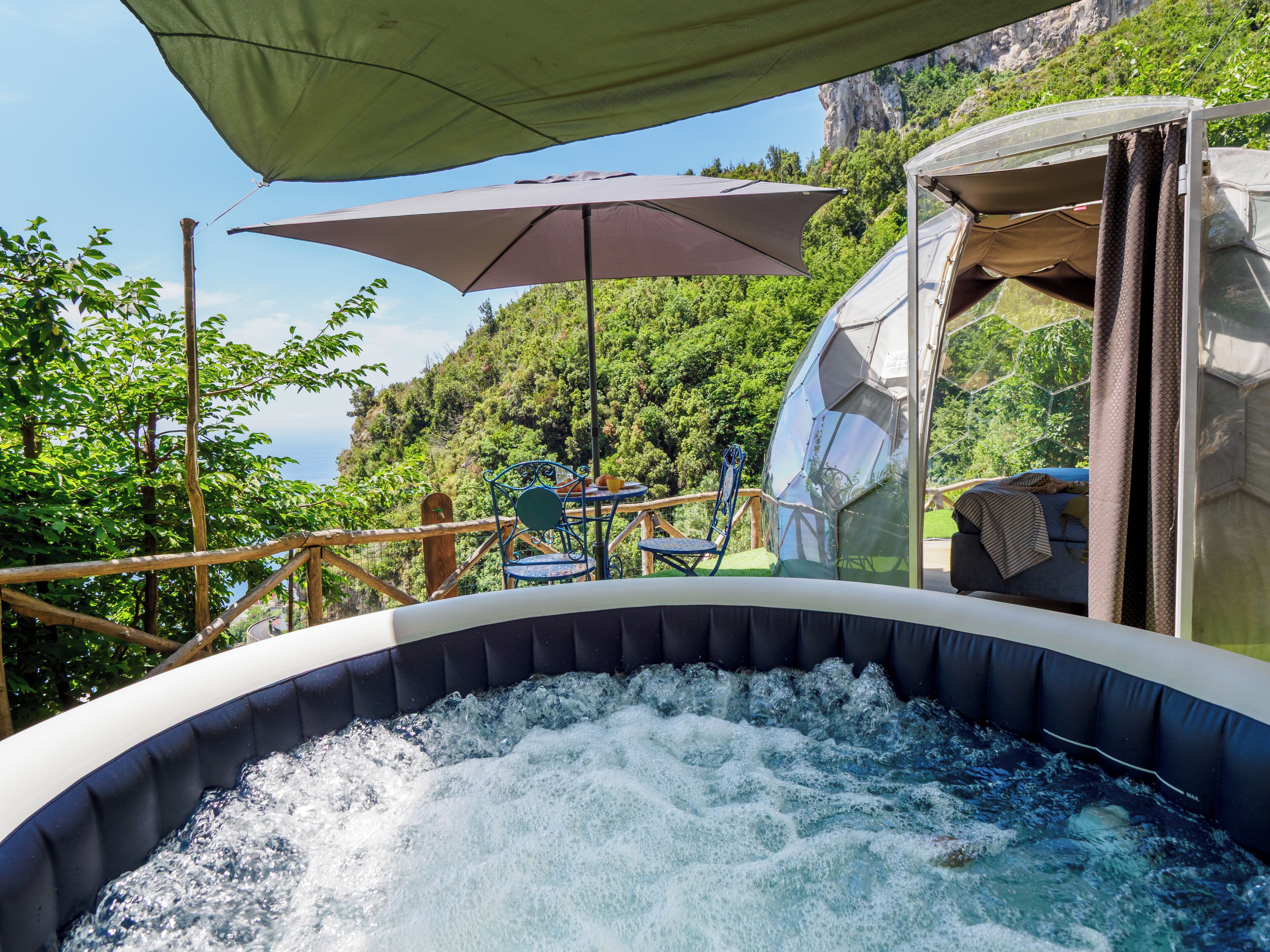 Un bain à remous extérieur offrant une vue sur un paysage montagneux, une terrasse en bois et une table avec des chaises.