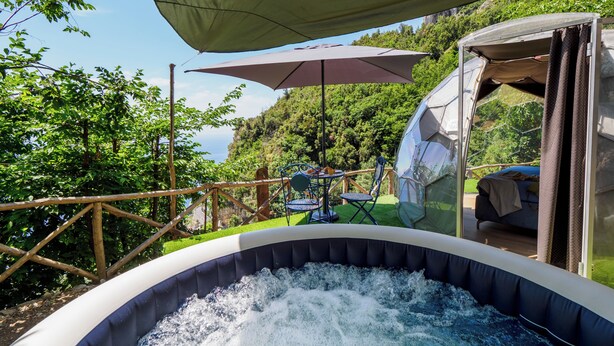 An outdoor hot tub with a view of a forested hillside, a dining area with chairs and an umbrella, and a geodesic dome structure. - De Vivo Realty - Boscosfera Amalfi (Amalfi)