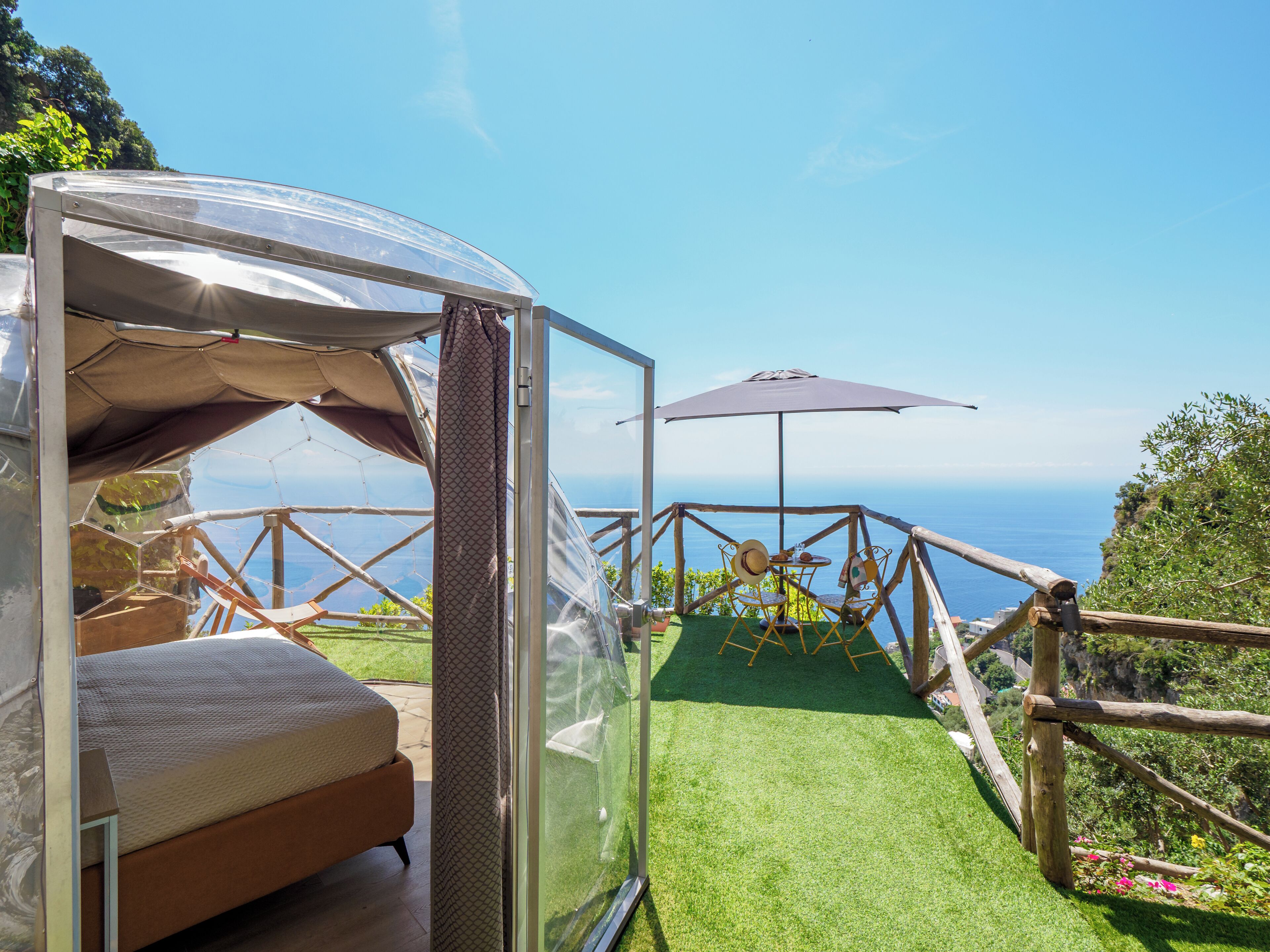 A transparent tent with a bed inside, a small table with chairs, and a view of the sea.