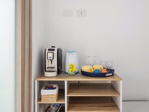 Une petite cuisine équipée d’une machine à café, d’une bouilloire, d’un panier de fruits et d’un panier en osier posé sur une étagère en bois.