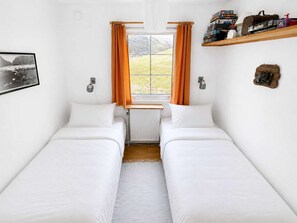 A room with two single beds, a radiator, a window with curtains, a shelf with books, and a framed picture on the wall. - Saksun Mountain & Beach Cottage (Saksun)