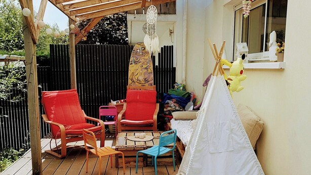 Une terrasse en bois avec des chaises rouges et bleues, une tente indienne blanche et une fenêtre donnant sur un beau paysage.